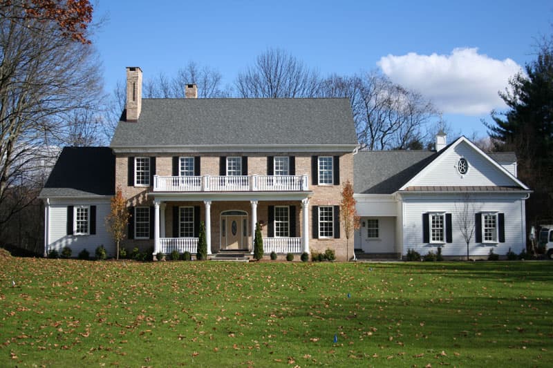 House plan exterior: Two-story brick and vinyl home with a central second-floor balcony, prominent front entrance, and attached gabled structure.