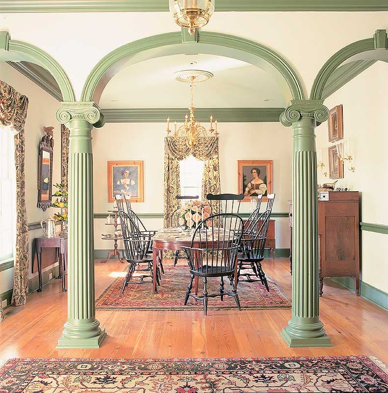 Dining room with fluted columns supporting arched openings, oval table, Windsor chairs, and oriental rugs.