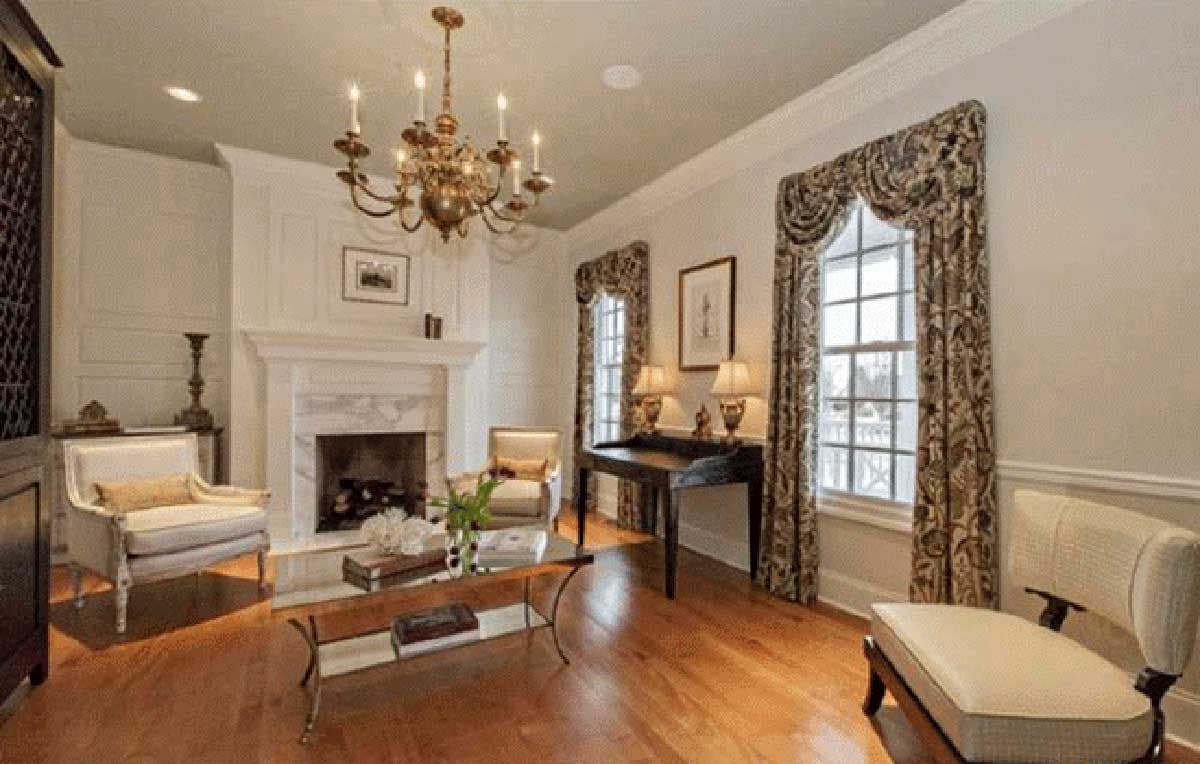 Living room with marble fireplace, ornate chandelier, and large windows with patterned drapes.