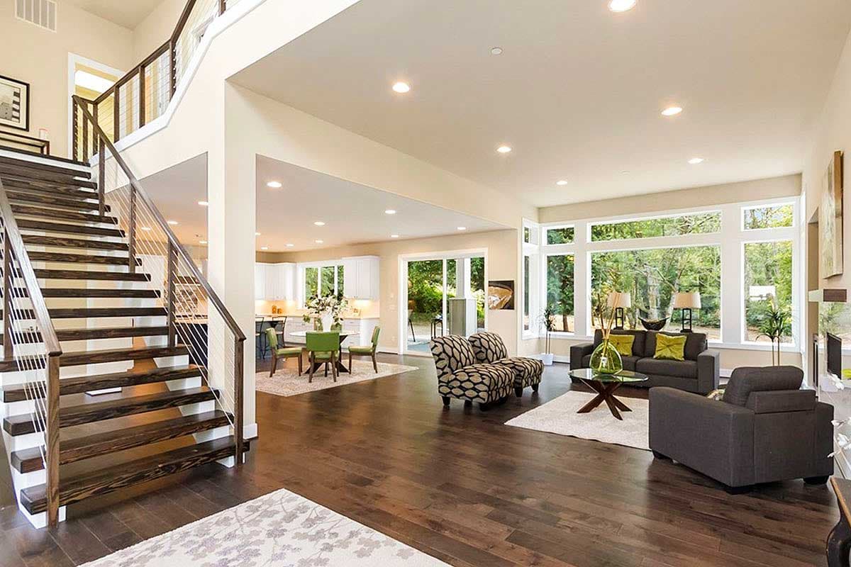 Open concept great room with modern staircase, dining area, and large windows overlooking wooded landscape.