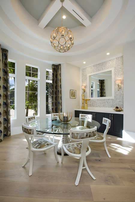 Dining room with glass table, upholstered chairs, built-in buffet, and tiered ceiling with decorative chandelier.
