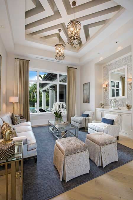 Living room with coffered ceiling, chandeliers, large windows overlooking patio and pool, and built-in cabinetry.
