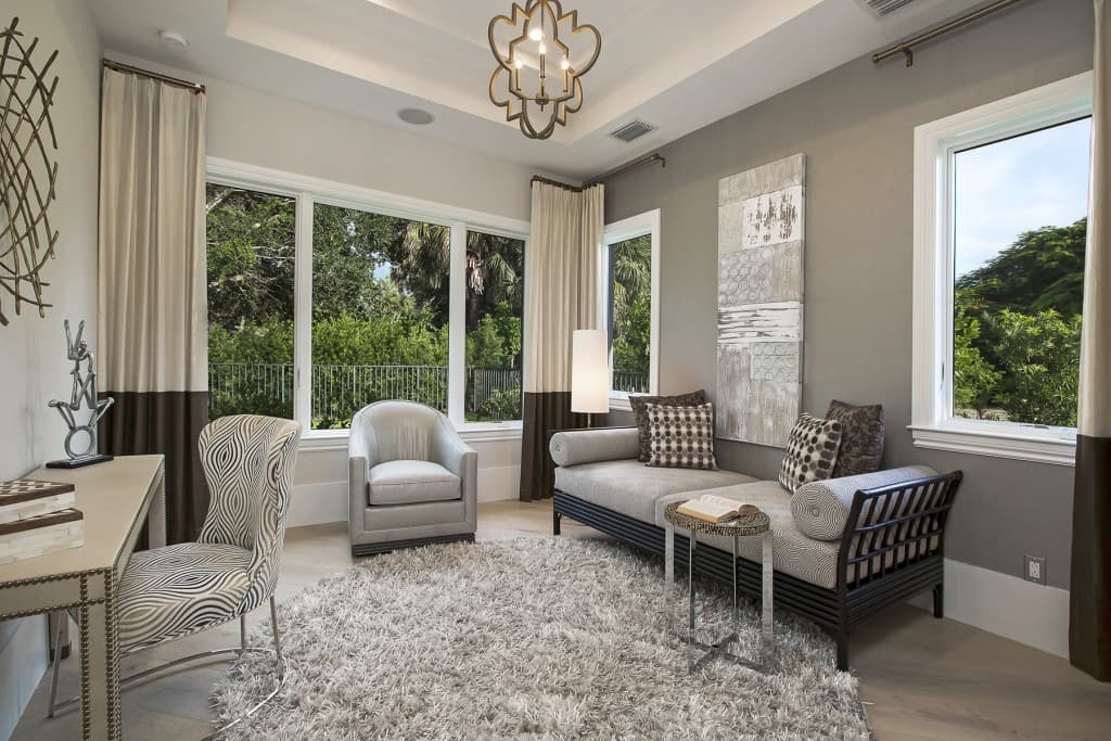 Interior view of a living room with a sofa, chaise, armchair, desk, and large windows overlooking greenery.
