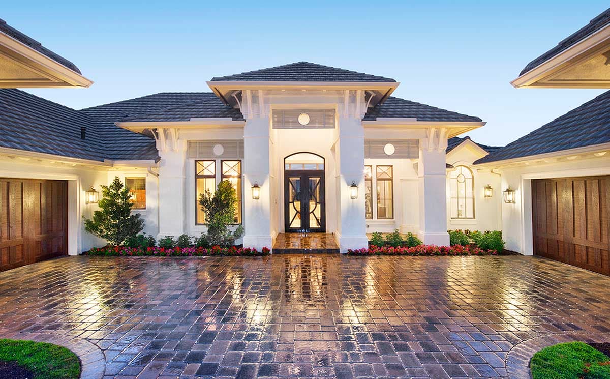 Elegant white home with dark roof and wooden garage doors, with a brick driveway reflecting the lights.