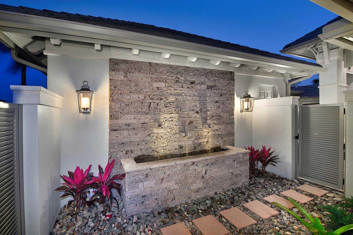 Exterior view of an upscale home with a stone wall fountain, lit by lanterns and accented with colorful plants.
