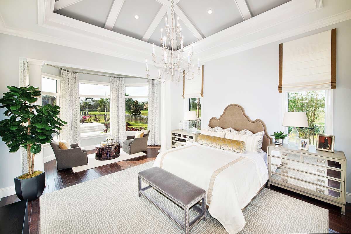 Luxury bedroom with a chandelier, large windows with a view, a white bed, and a gray bench at the foot of the bed.