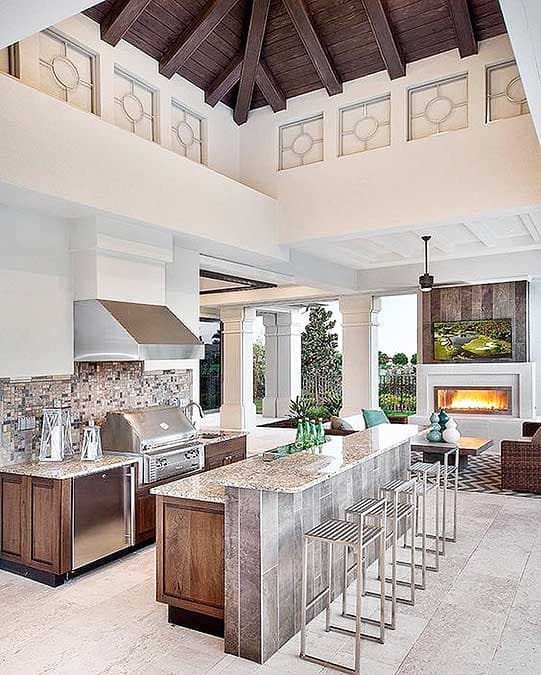 Outdoor kitchen with grill, island with bar stools, and fireplace. High ceiling with wooden beams and decorative windows.