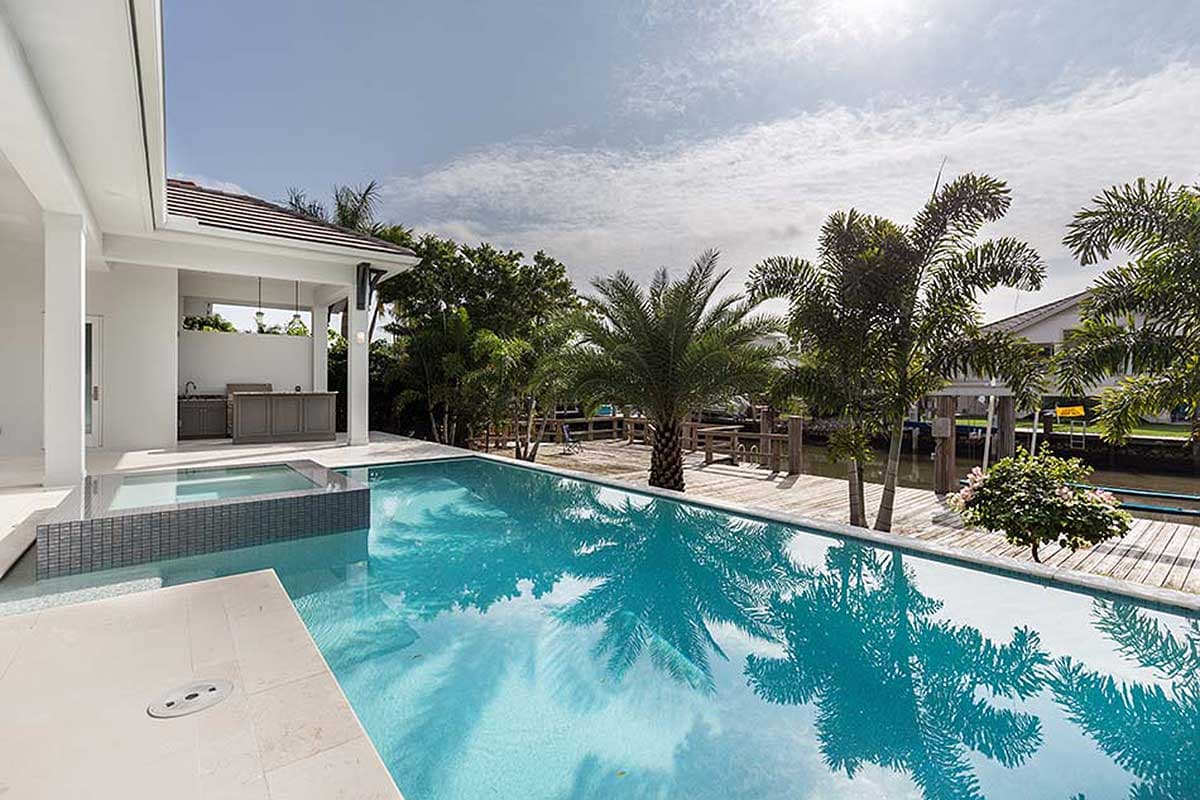 Modern house exterior with covered outdoor kitchen, infinity pool, and canal-side wooden deck.