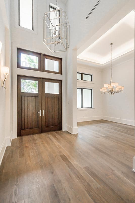 Interior view of house entry with double wooden doors, glass transoms, high ceilings, and decorative chandeliers.