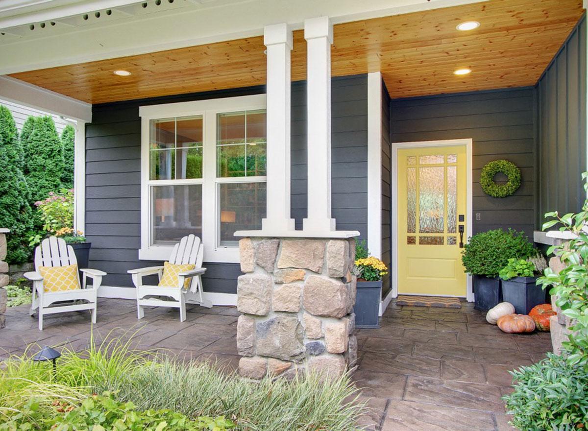 House plan exterior: Dark siding, stone accent pillar, covered porch with wood ceiling, two white chairs, and a yellow door.