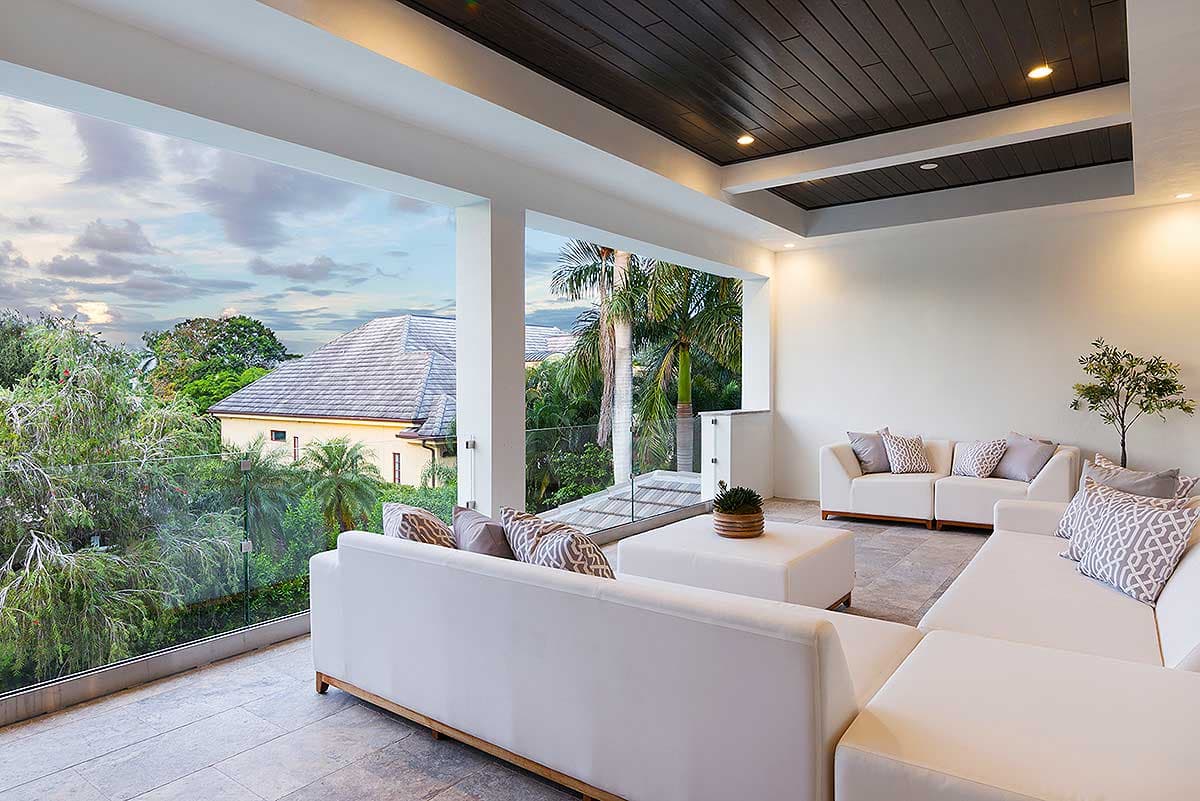 Covered patio with white sectional sofas, glass railings, and a dark wood paneled ceiling. Overlooks lush greenery and a distant house.