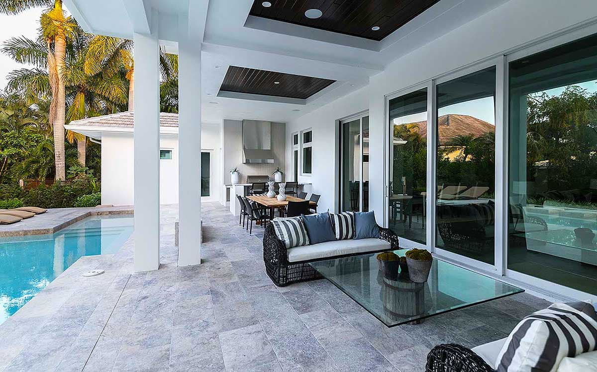 Covered outdoor living space with pool, seating area, and outdoor kitchen, featuring large sliding glass doors and wood-paneled ceiling.