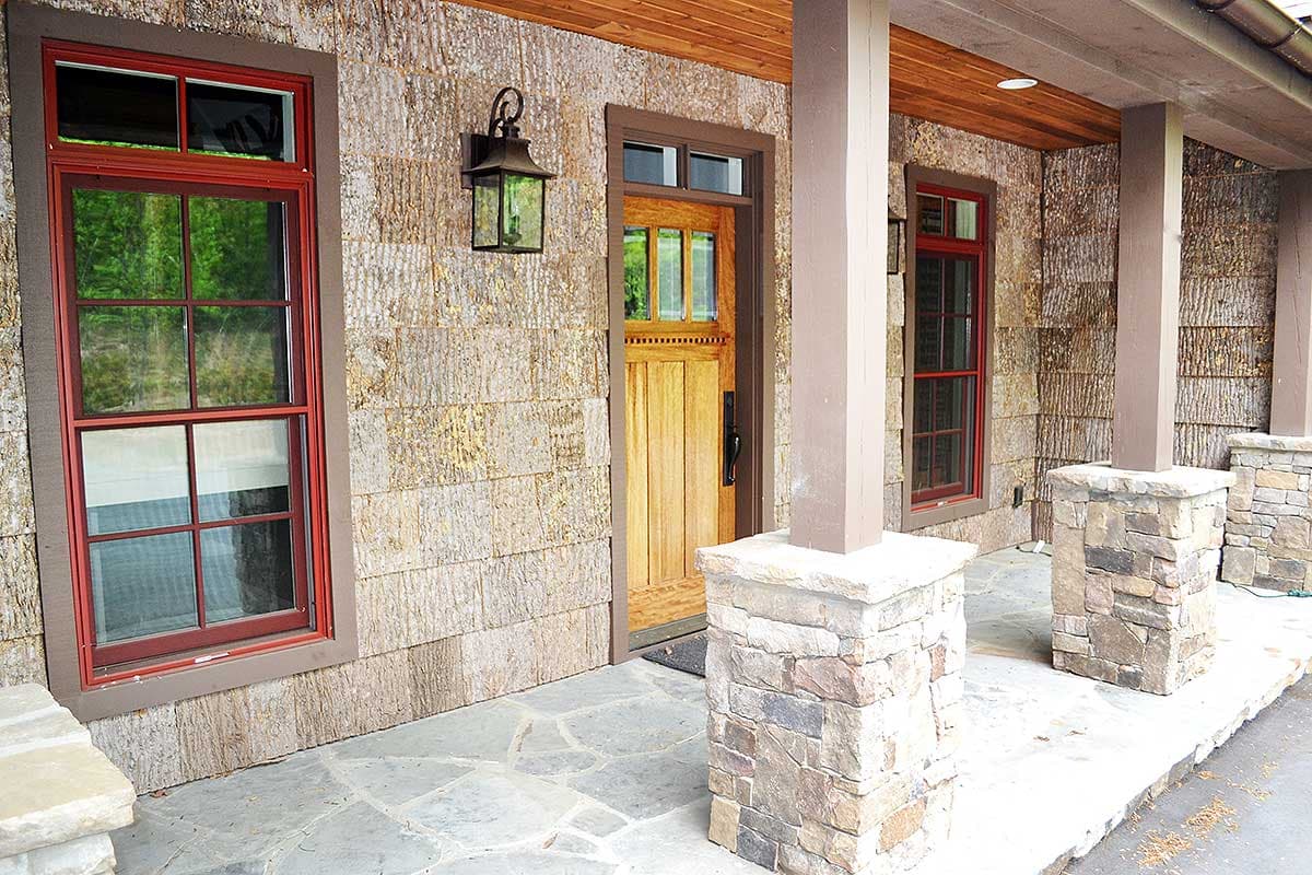 House plan exterior with wood shake siding, stone pillars, red-trimmed windows, and a wooden front door with glass panes.