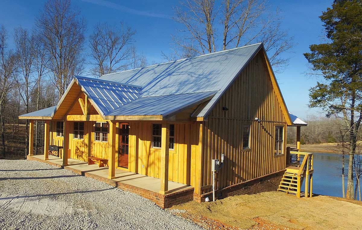 Log cabin house plan exterior with metal roof, wraparound porch, and lakeside setting.