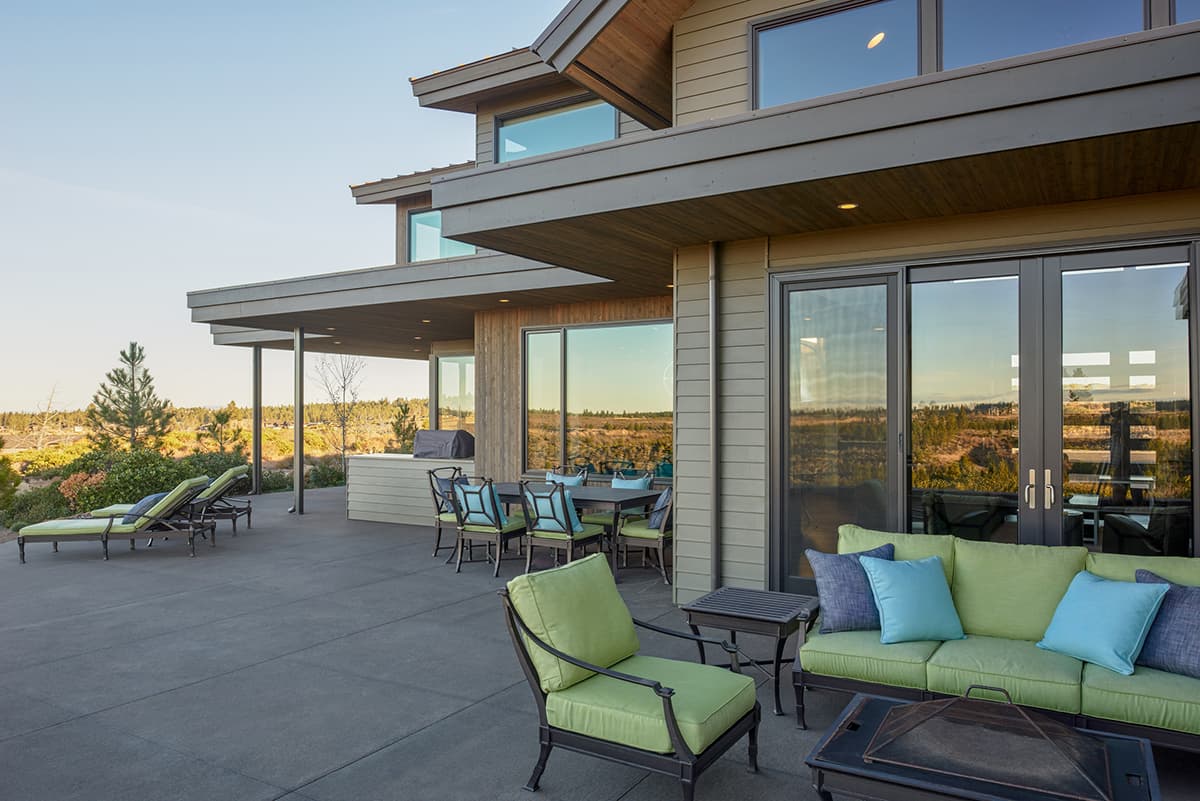 Modern house exterior with layered rooflines, horizontal siding, large windows, and covered patio with outdoor dining and lounge areas.