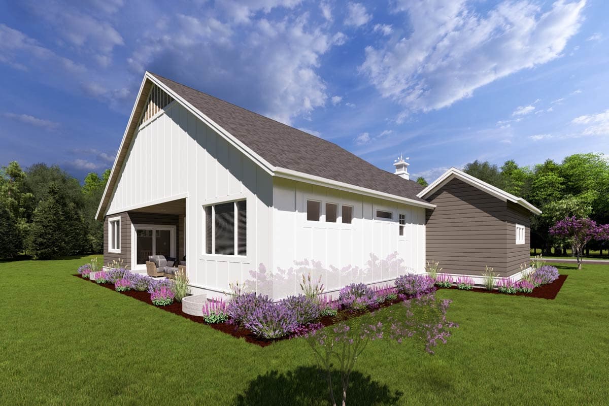 A modern farmhouse with white siding and a dark brown roof. The house features a porch and is surrounded by a green lawn and flowering plants.