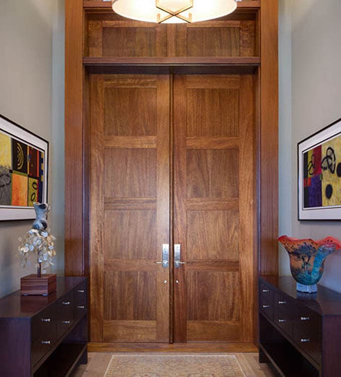 Double wooden doors with paneling flank a hallway featuring console tables and artwork. Modern interior.