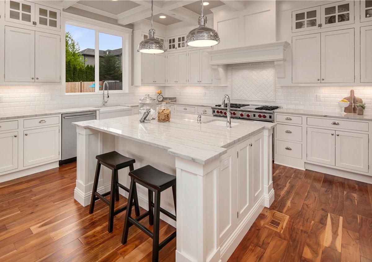 Kitchen interior with white cabinets, marble island with seating, stainless steel appliances, subway tile backsplash, and hardwood floors.