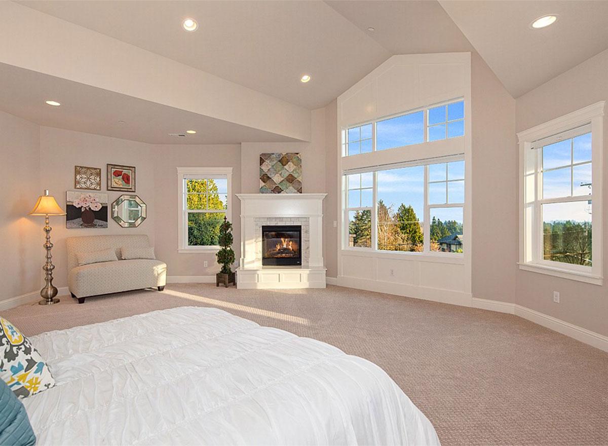 Bedroom with fireplace, vaulted ceiling, and large grid windows overlooking trees and landscape.