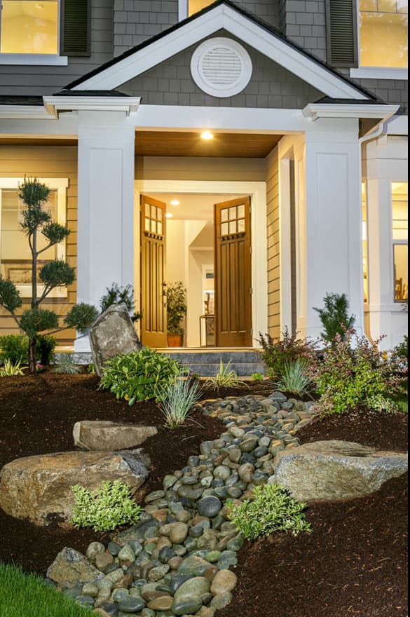 House plan exterior featuring double entry doors, white columns, a peaked gable with circular vent, and a dry creek bed landscape.