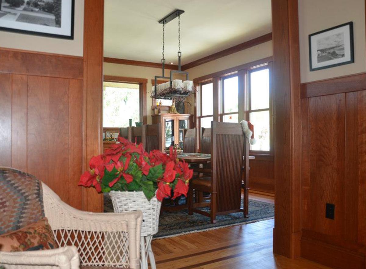 Dining room with wooden table, chairs, buffet, and large windows. Chandelier hangs overhead. Wood wainscoting visible.