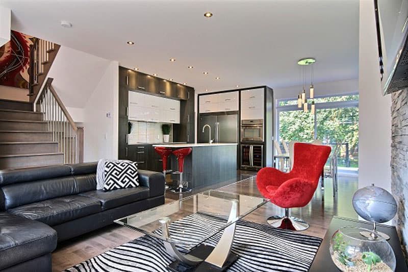 Open concept living space with modern kitchen, island, black leather sofa, glass coffee table, and red accent chair.