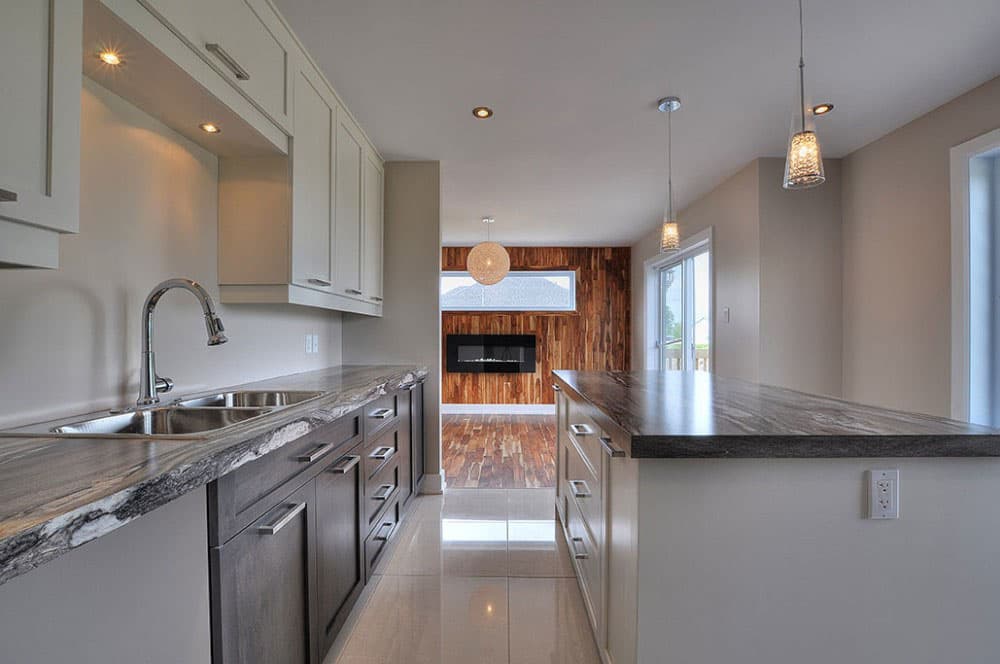 Kitchen with island, dual sink, dark lower cabinets, and view of living area with fireplace and wood accent wall.