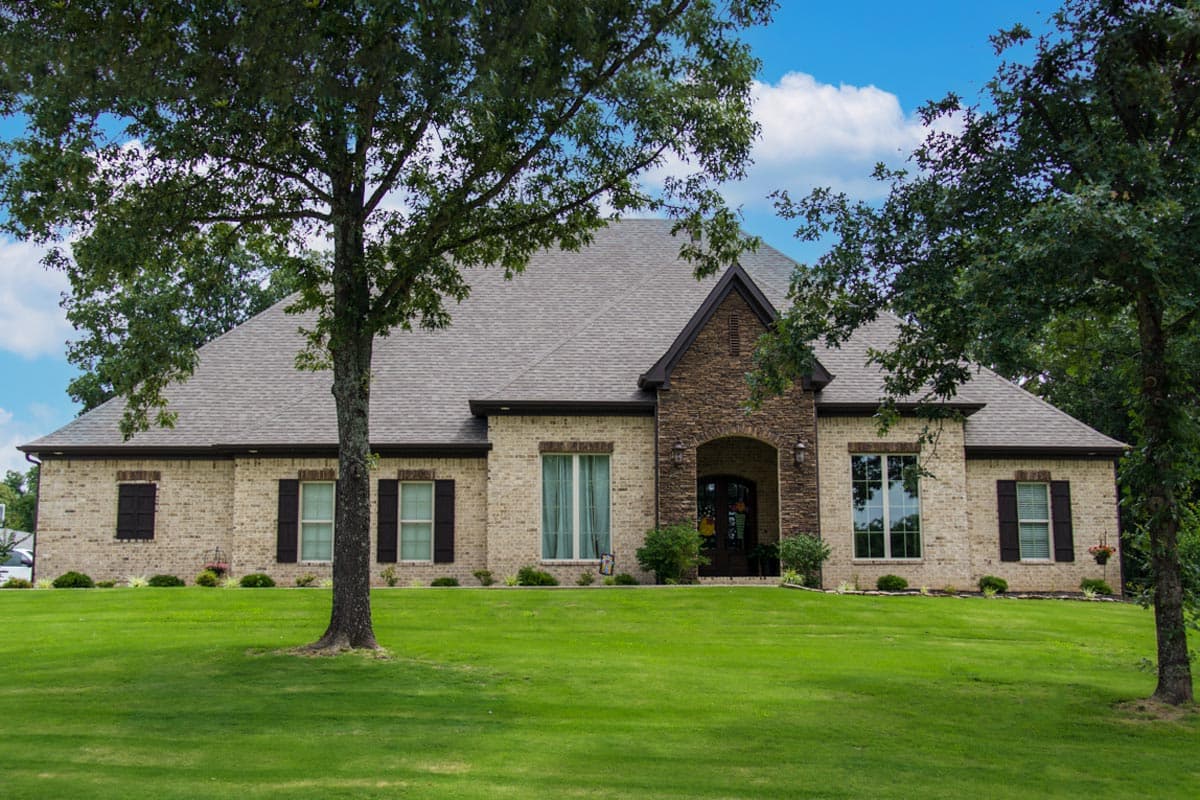 House plan exterior of a two-story brick home with a gabled stone entry, dark shutters, and a prominent hipped roofline.