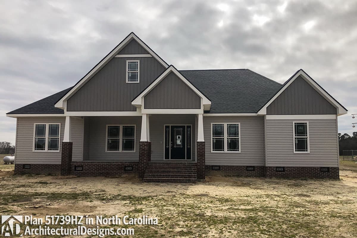 House plan exterior: Ranch style, single story with gabled dormers, covered porch supported by brick columns, and vertical siding.