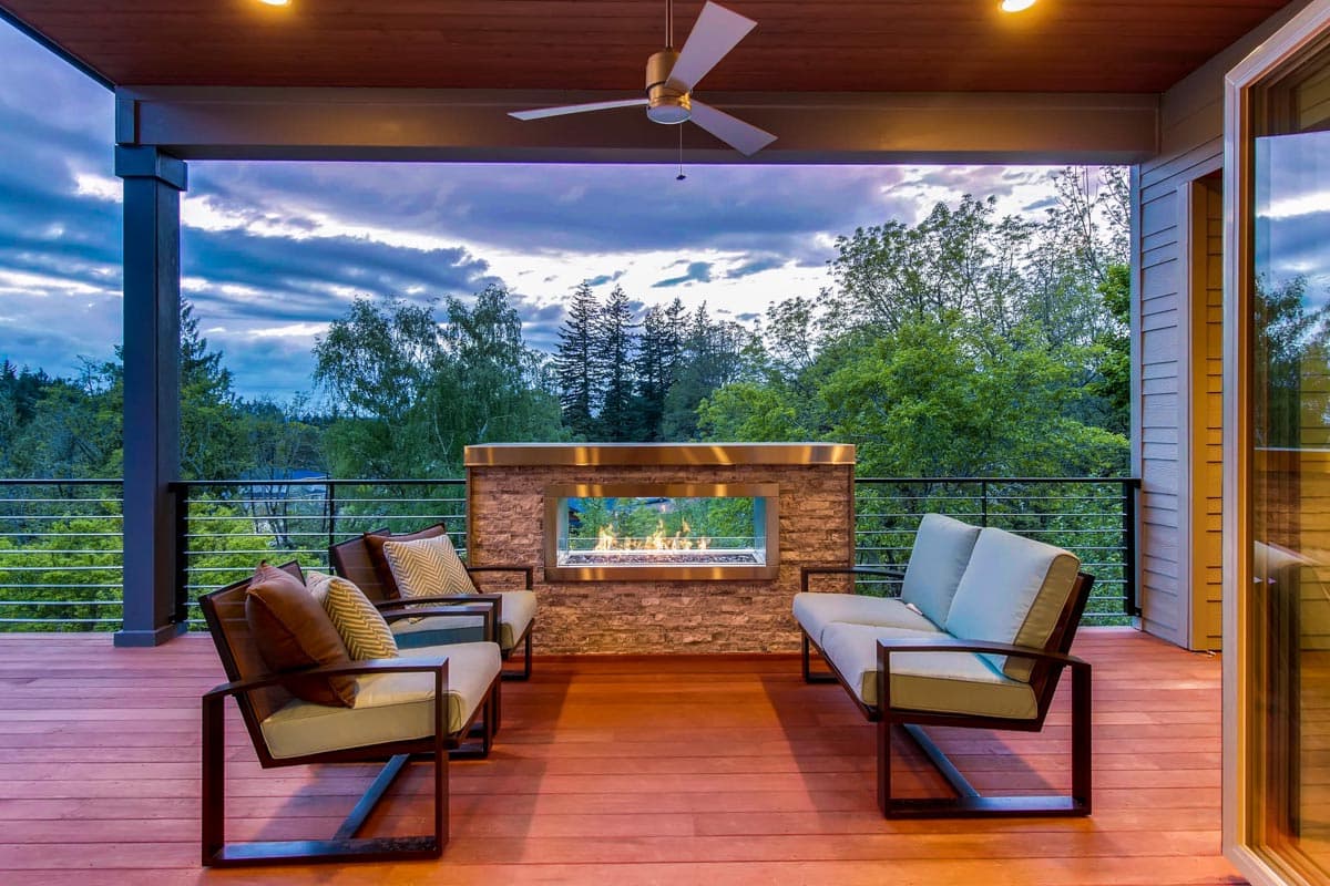 Covered deck with wood flooring, contemporary seating, and linear gas fireplace. House plan exterior.