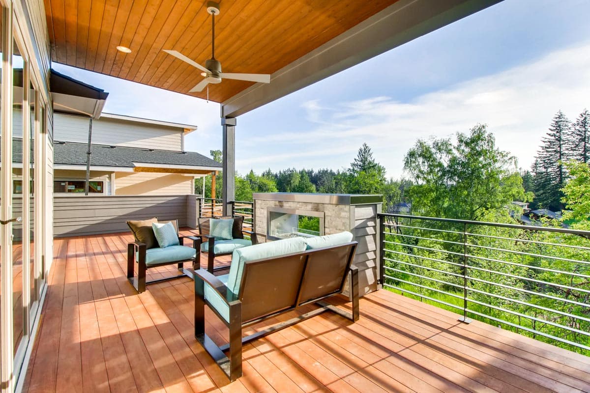 Deck with seating, modern outdoor fireplace, and wooded views. Features wood ceiling and fan.