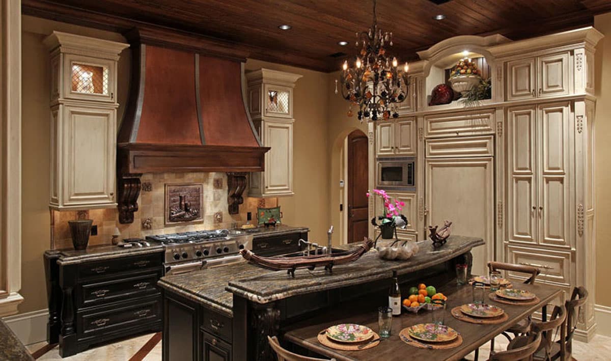 Kitchen interior with ornate cabinetry, a large island with seating, copper range hood, and a decorative chandelier.