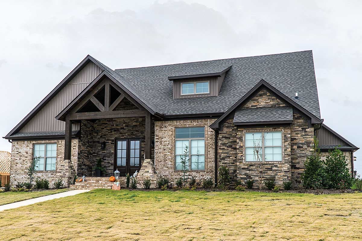 House plan exterior with stone and brick facade, gabled roof, and timber-framed porch entry. Includes dormer and multi-pane windows.