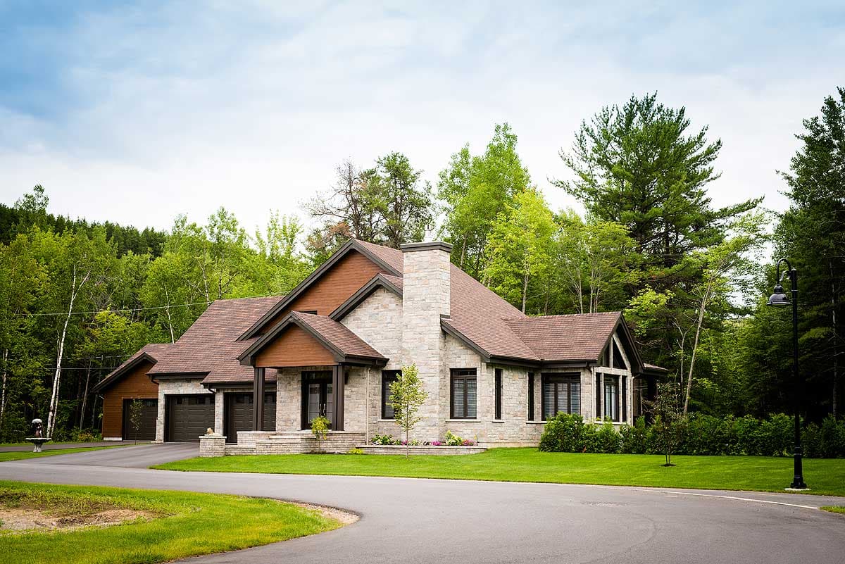A large, modern home with a stone facade and a brown roof. The house is surrounded by green grass and tall trees, with a curved driveway.