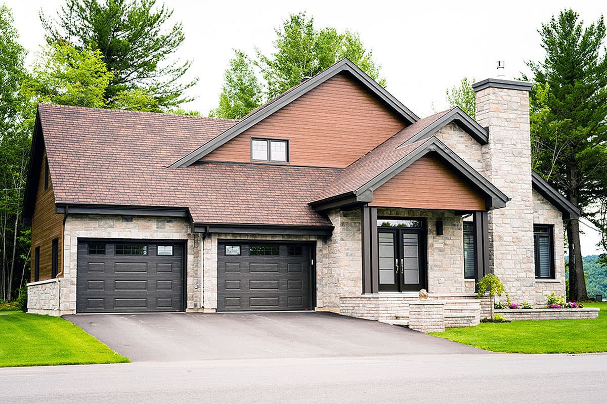 A modern, stone-clad house featuring a brown roof, two-car garage, and a brick chimney. Trees surround the house on a bright day.