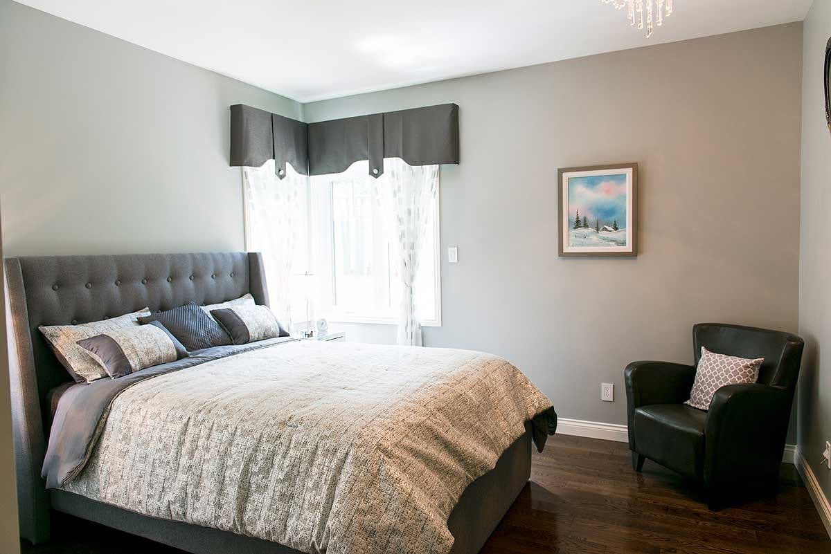 A bedroom with a gray bed, a black armchair, and a framed painting on the wall. The room features a window with curtains and dark wood flooring.