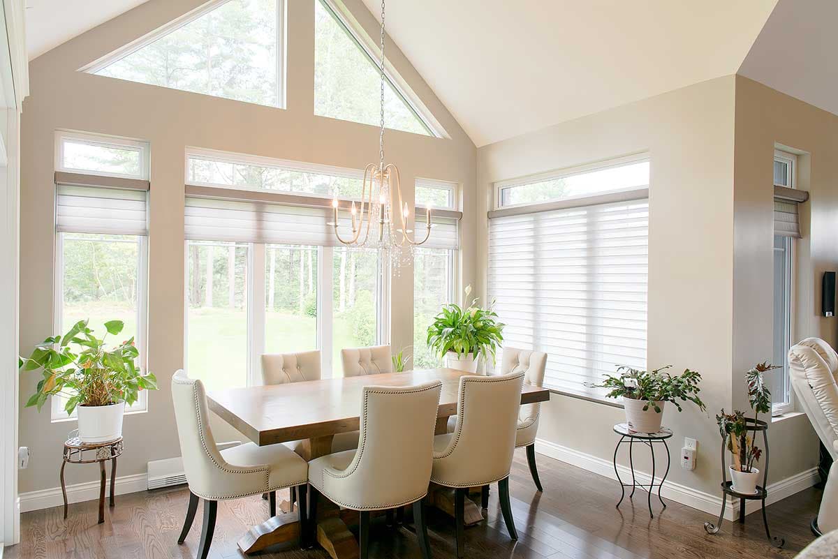 Spacious dining room with large windows overlooking trees. A wooden table is surrounded by six beige upholstered chairs, and a chandelier hangs above.
