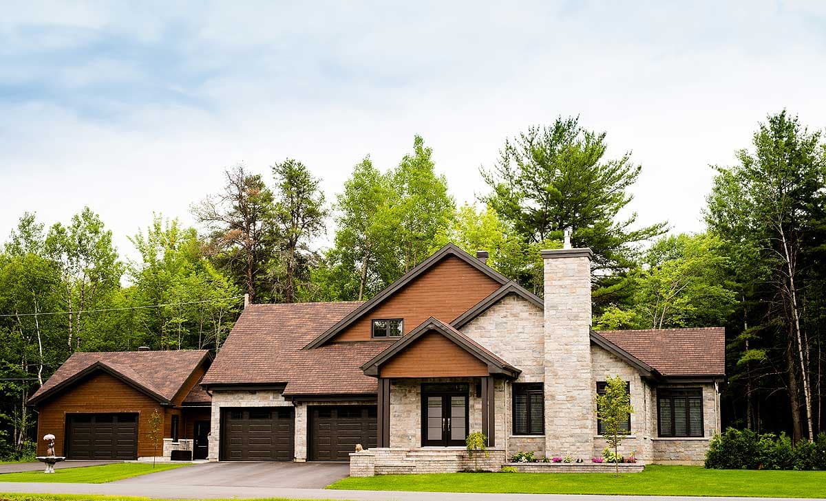 A large, two-story house with a stone and wood exterior sits on a lush green lawn, surrounded by tall trees under a cloudy sky.