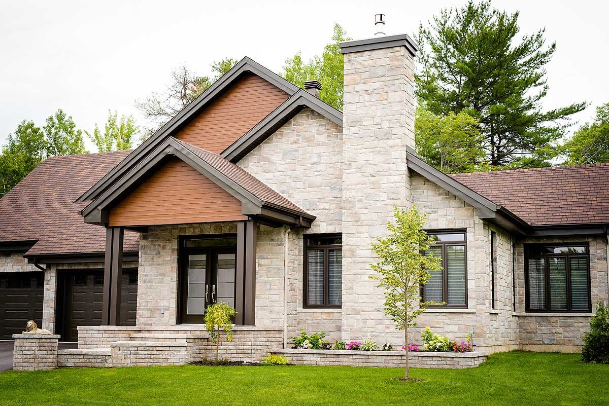 A modern, single-story house featuring a stone facade and brown roof. A large chimney stands next to a gabled roof. Green lawn and trees surround the building.