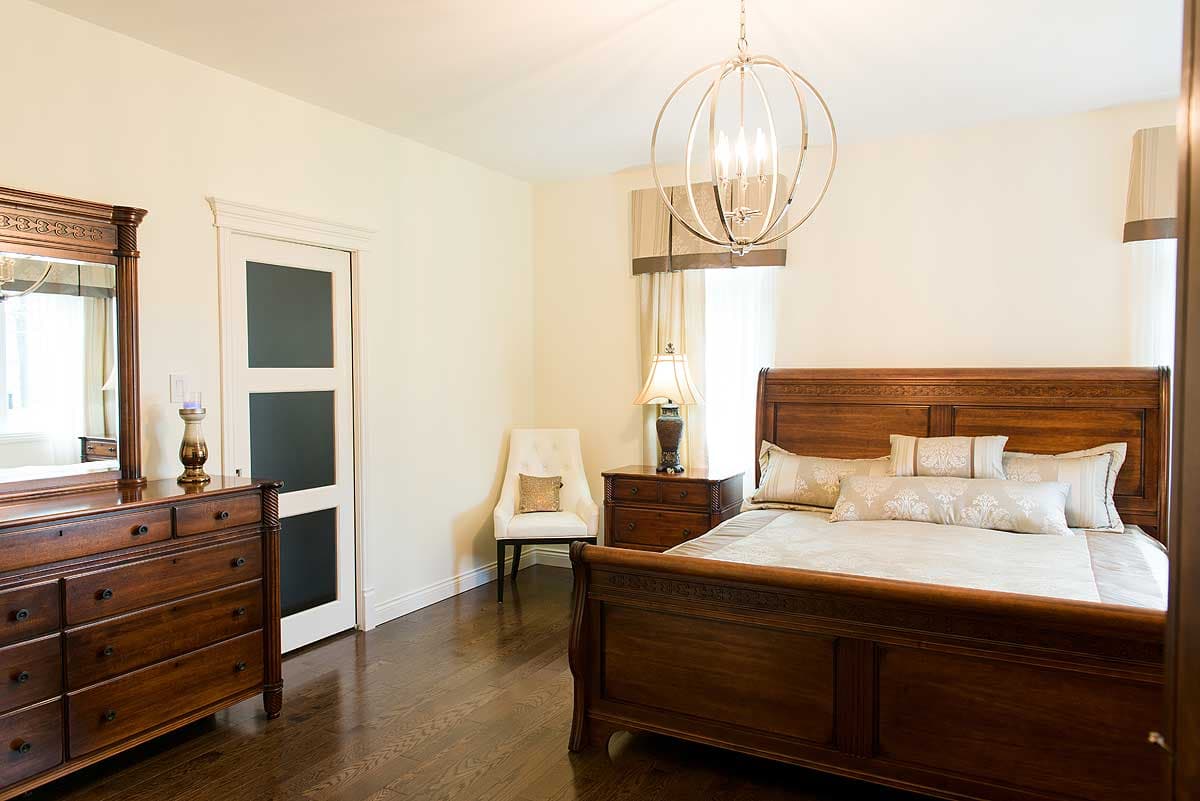 A bedroom features a large, dark wooden bed, dresser, and nightstand. A chandelier hangs above the bed, with a white chair and a door visible.