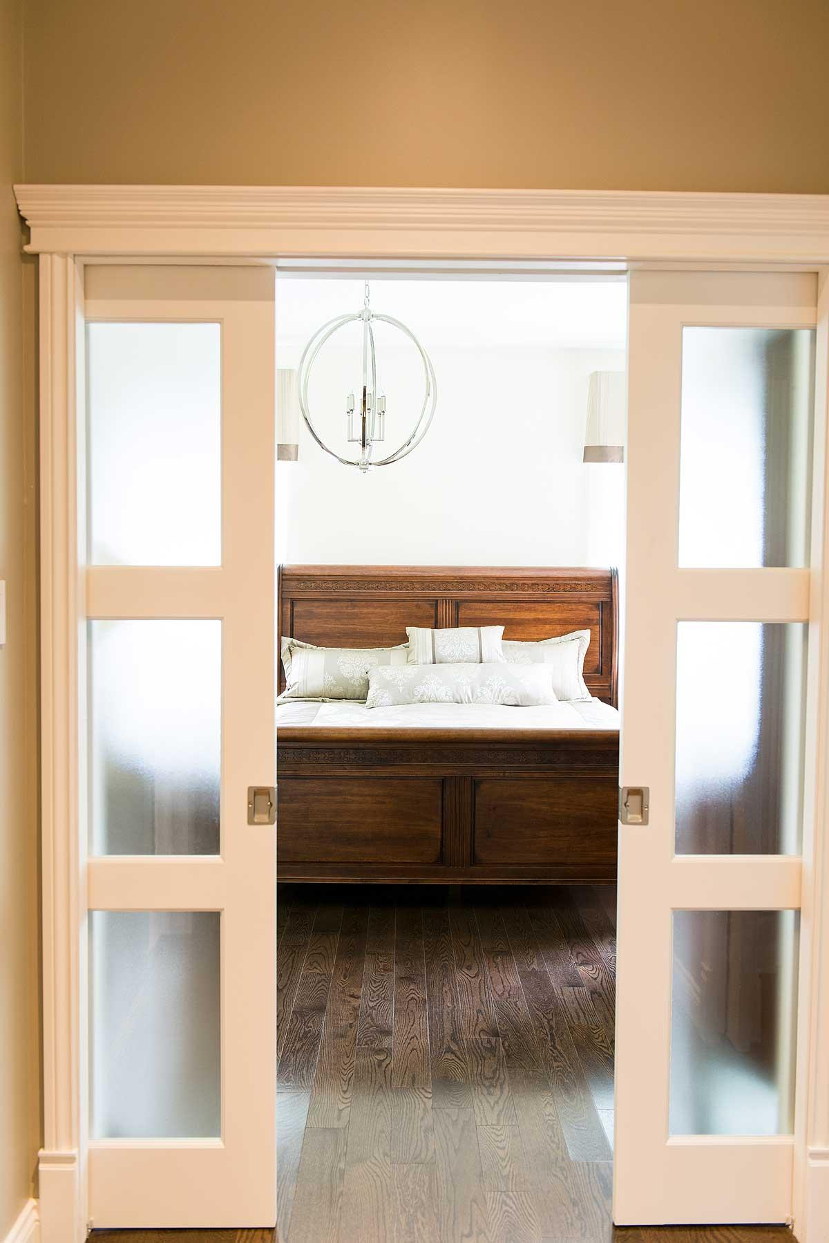 A bedroom is viewed through open frosted glass doors. A large, ornate wooden bed is the focal point, with a decorative chandelier hanging above.