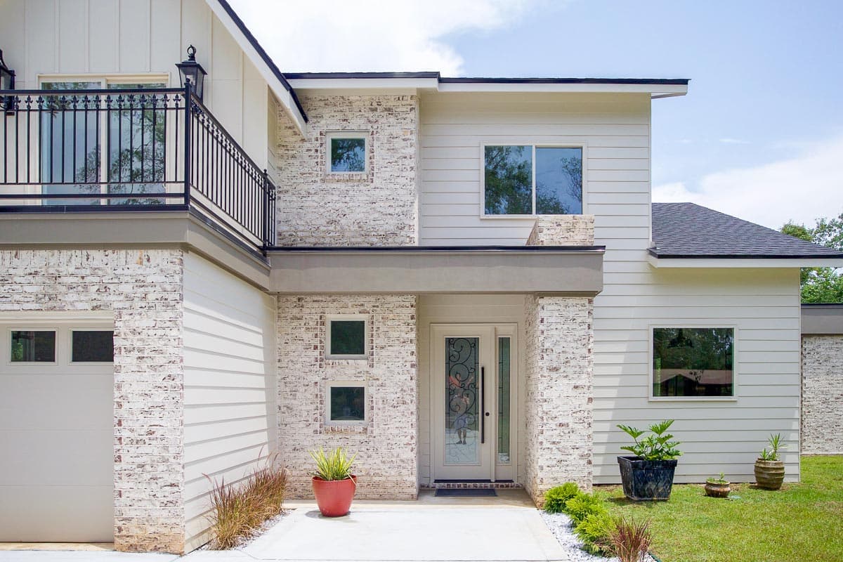 Modern house plan exterior with white brick, vertical siding, multiple windows, a second-story balcony with black railing, and a garage.