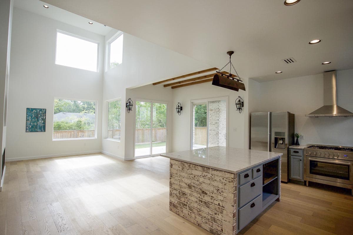 Open concept living area with vaulted ceiling, large windows, wood beams, kitchen island with brick base, and stainless steel appliances.