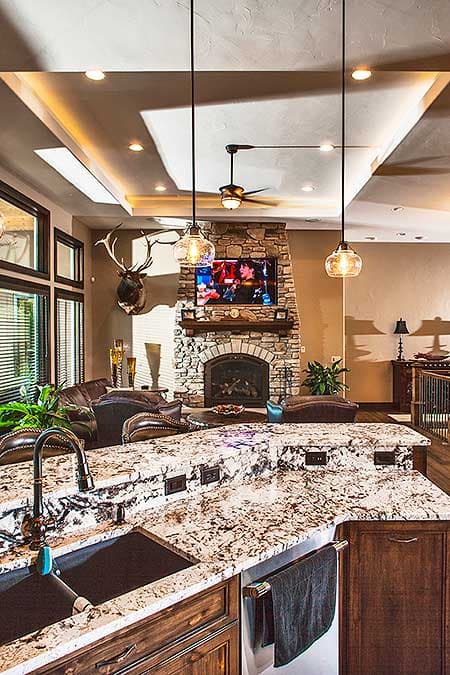 Kitchen island with granite countertop, double sink, and wood cabinets; adjacent great room with stone fireplace and television.