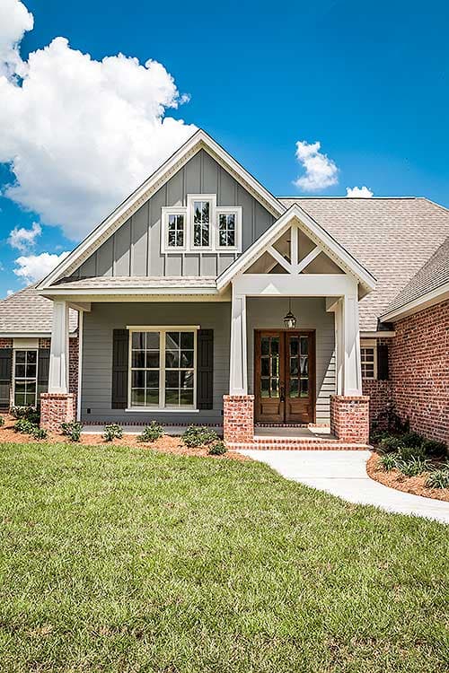 House plan exterior: Modern Farmhouse style with a covered porch, brick piers, double front doors, and triple gable dormer.
