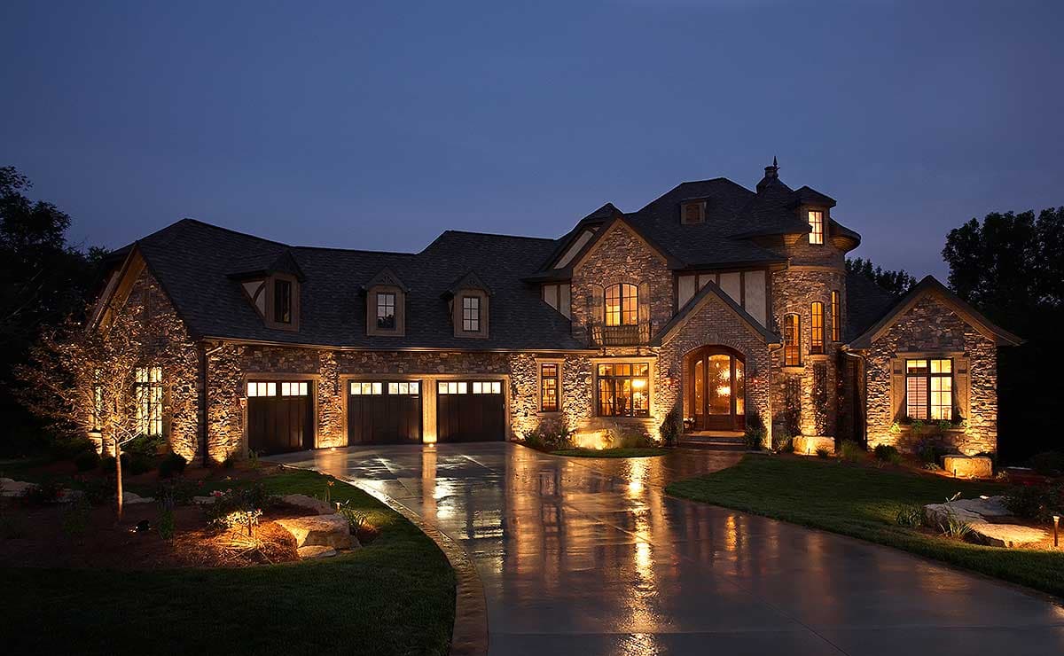 House plan exterior with stone facade, multi-car garage, and multiple gables. Features a turret and a prominent front entry, with night lighting.