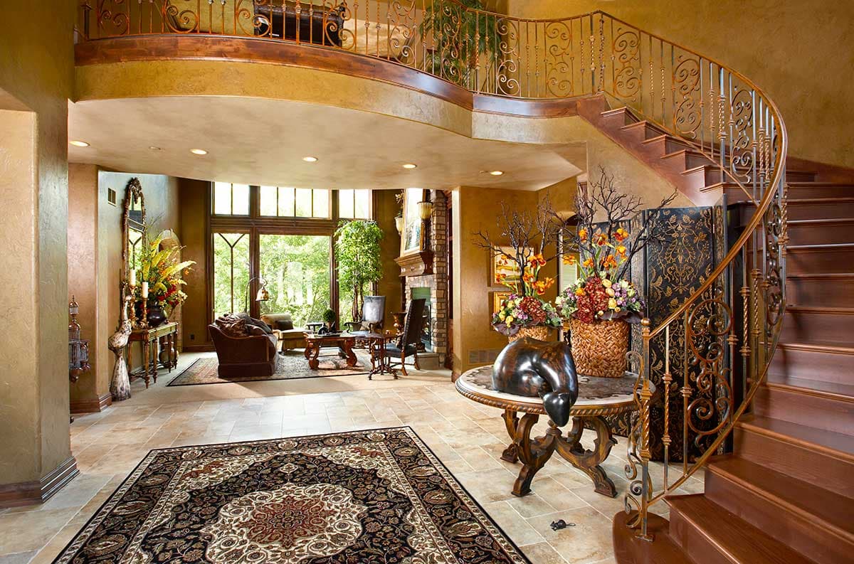 Interior view of a grand foyer with a curved staircase, wrought iron railings, large windows, and a sitting area.