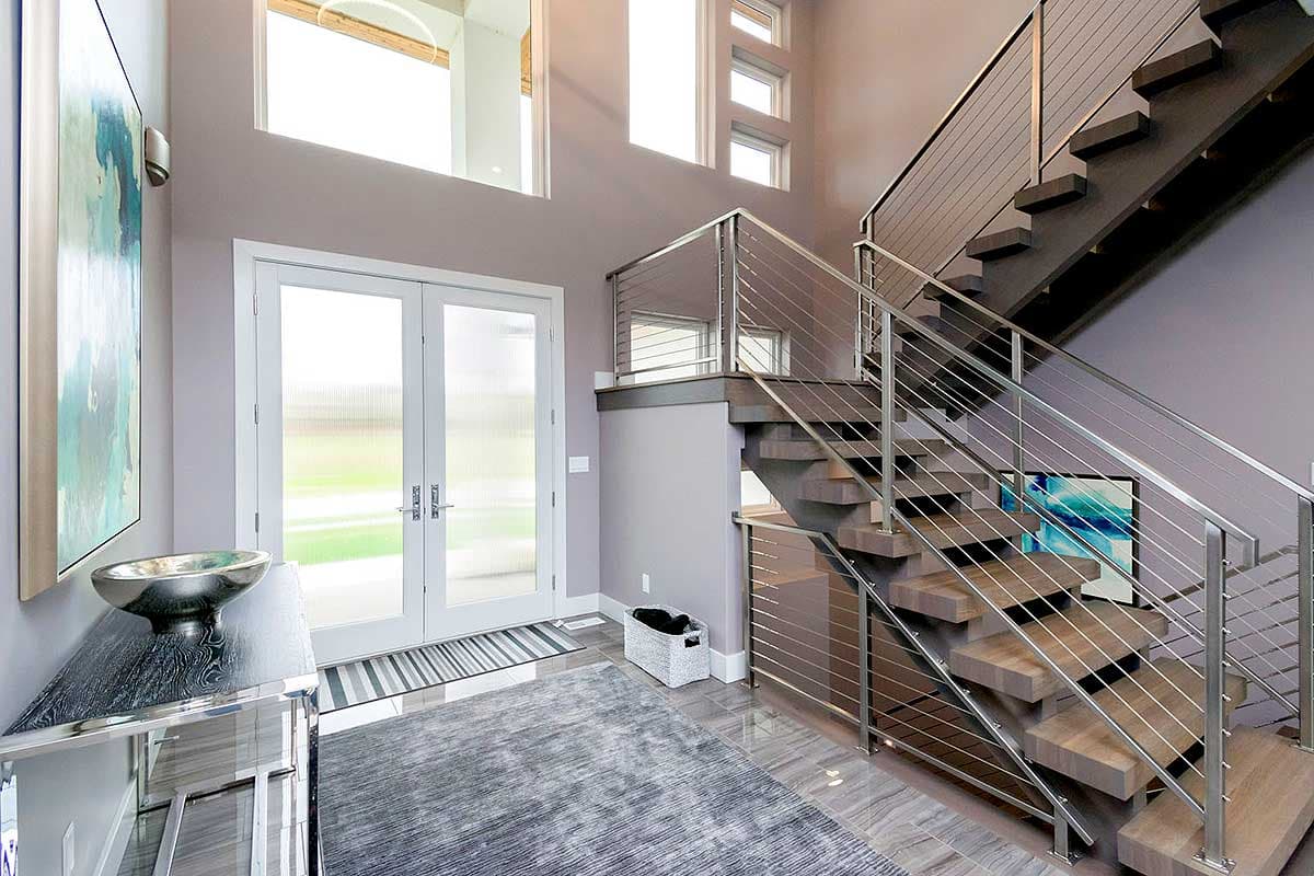 Interior view of a modern house foyer with a floating staircase, double glass doors, and high windows.