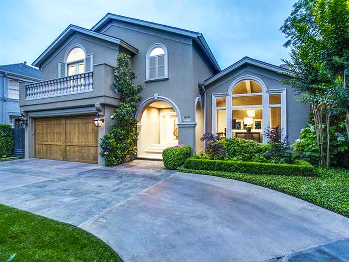 House plan exterior with stucco facade, arched entryway and window, garage, balcony, and gabled roof. Landscaping includes hedges and lawn.