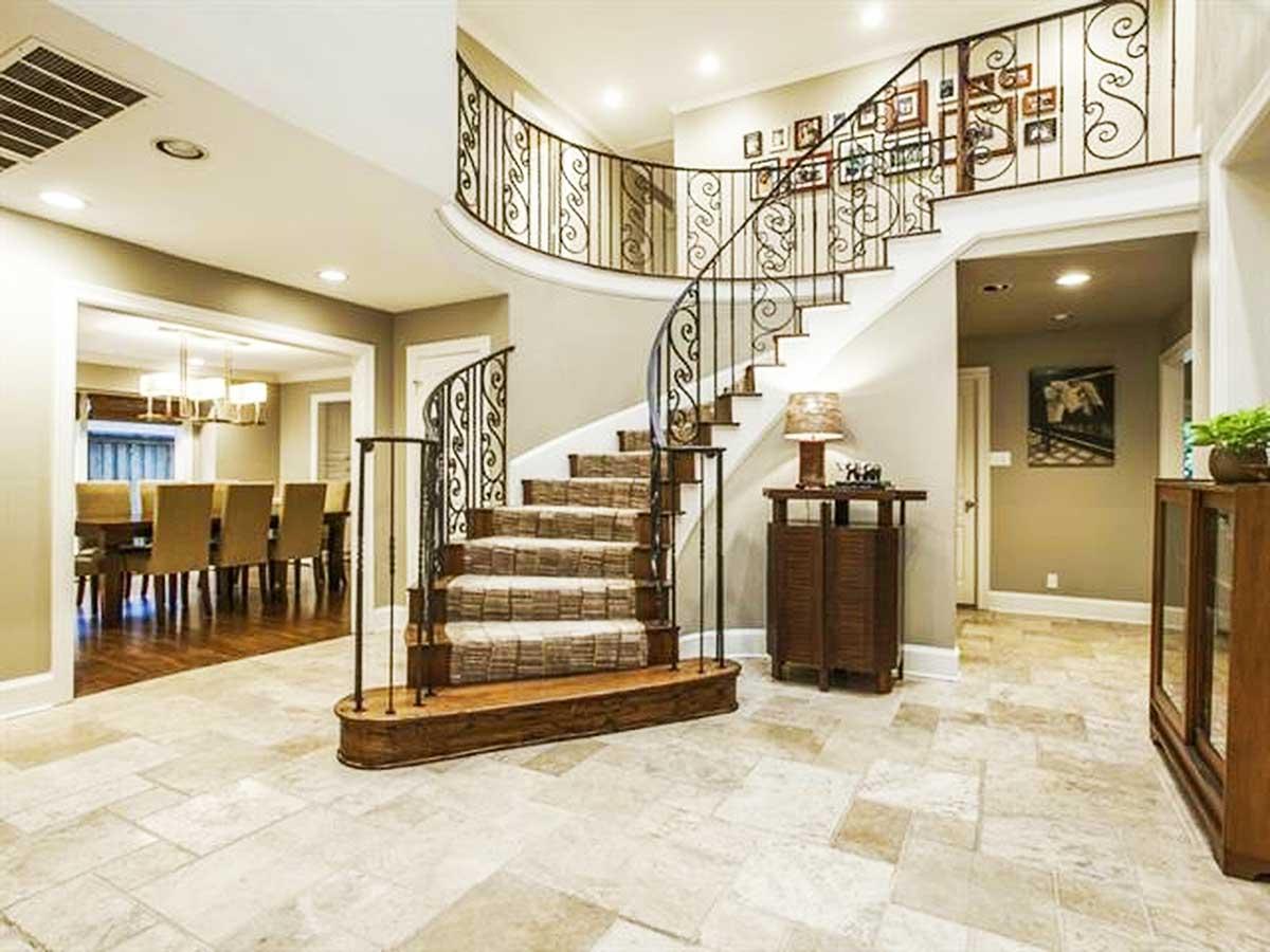 Interior view of a foyer with a sweeping curved staircase featuring ornate metal railings, leading to an upper landing with framed photos.